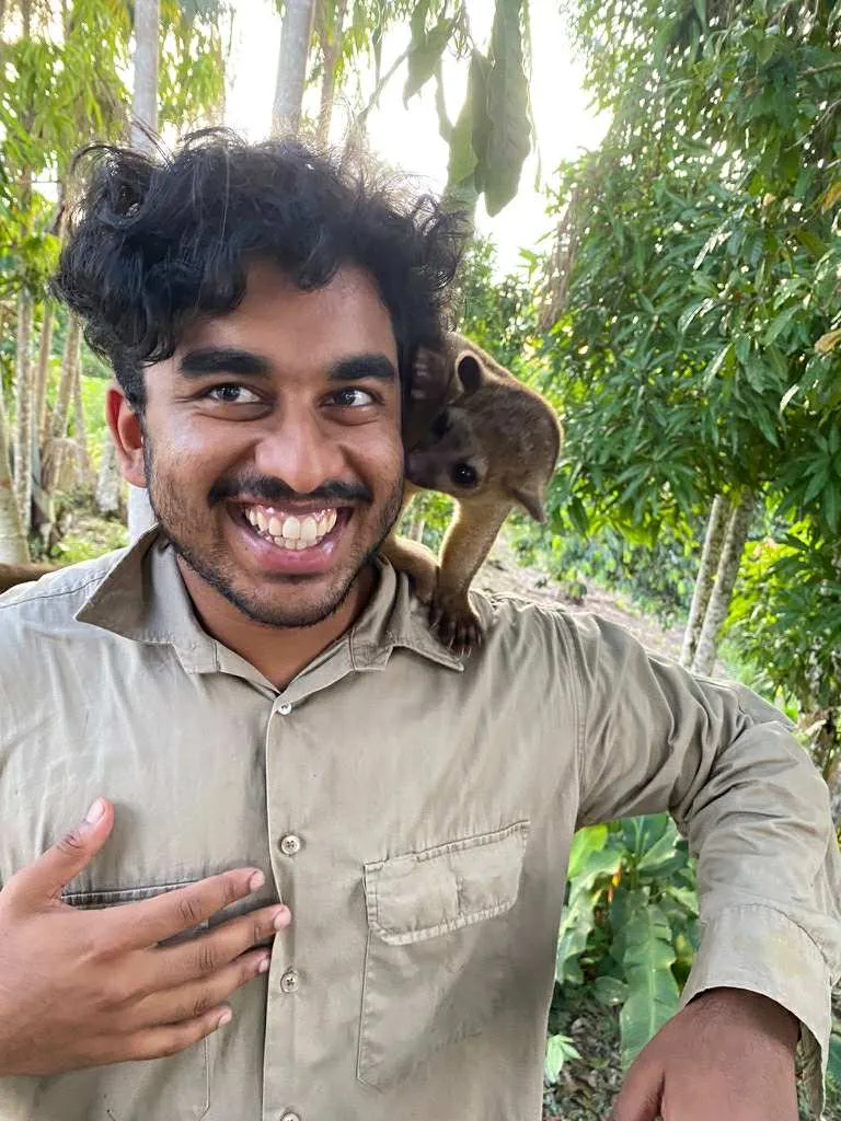 Niraj Meisuria smiling in a rainforest clearing, with a small furry mammal perched on his shoulder